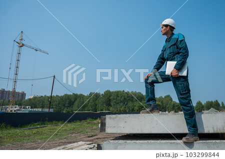 Caucasian male builder in hardhat holding laptop at construction site.  103299844