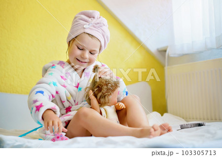 Cute smiling little girl child playing with her doll at home. Preschool child in bathrobe after taking bath brushing hairs of toy doll. Happy kid. 103305713