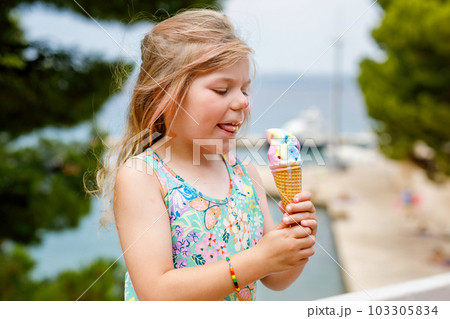 Happy preschool girl eating colorful ice cream in waffle cone on sunny summer day. Little toddler child eat icecream dessert. Sweet food on hot warm summertime days. Bright light, colorful ice-cream 103305834