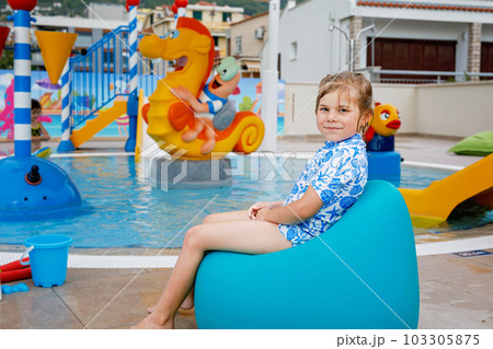 Little preschool girl sitting by outdoor swimming pool in hotel resort. Child learning to swim in outdoor pool, splashing with water, enjoying, laughing and having fun. Family vacations. Little preschool girl sitting by outdoor swimming pool in hotel resort. Child learning to swim in outdoor pool, splashing with water, enjoying, laughing and having fun. Family vacations. 103305875
