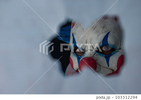 Creepy clown face peering through a hole in the partition. The face of a man wearing a creepy clown mask peering at his victim through a hole. A clown madman. 103312294