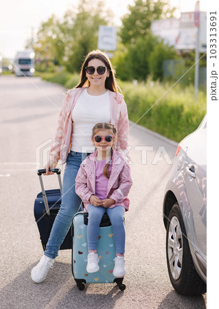 Adorable little girl sits on her kid suitcase and waiting for the trip with her mom. Two beautiful girls prepare to journey. A car is parked on the side 103313691