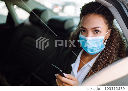 Woman in a sterile mask in taxi car on a back seat looking out of window checking her cell phone. 103313879