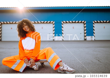 Beautiful African American woman posing with a skateboard. Hipster woman with curly hair with skateboard at sunset. 103314639