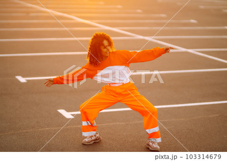 Young African American woman - dancer dancing in the street at sunset. Stylish woman with curly hair in an orange suit showing some moves. 103314679
