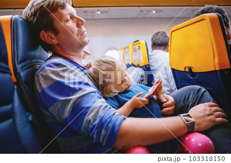 Young father holding his baby toddler daughter during flight on airplane going on vacations. Tired man and cute girl during night flight. child playing with smartphone. Family sitting in plane 103318150