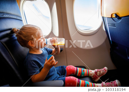 Adorable little girl traveling by an airplane. Small toddler child drinking orange juice sitting near aircraft window. Traveling abroad with kids. Family on summer vacations. Adorable little girl traveling by an airplane. Small toddler child drinking orange juice sitting near aircraft window. Traveling abroad with kids. Family on summer vacations. 103318194