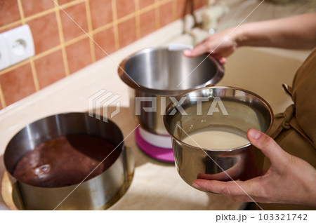 Close-up of housewife hands holding bowl with fresh milk or cream above a kitchen countertop, preparing whipped sweet dessert in the home kitchen interior. Culinary. Cuisine. Confectionery Close-up of housewife hands holding bowl with fresh milk or cream above a kitchen countertop, preparing whipped sweet dessert in the home kitchen interior. Culinary. Cuisine. Confectionery 103321742