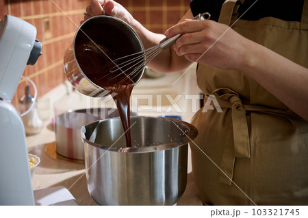 Close-up pastry chef pouring melted chocolate into a metal bowl of planetary mixer, mixing it with cream for preparing delicious chocolate cream or with whipped egg whites for making sponge biscuit Close-up pastry chef pouring melted chocolate into a metal bowl of planetary mixer, mixing it with cream for preparing delicious chocolate cream or with whipped egg whites for making sponge biscuit 103321745