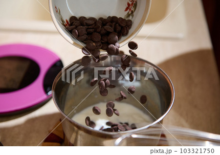 Close-up chips of organic dark belgian chocolate spilling out from a ceramic bowl into a metal pan. Melting chocolate. Preparing cream, candies or glaze. Still life Selective focus 103321750