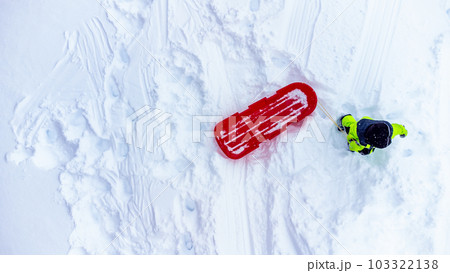 Aerial overhead drone photo of a child walking up a snow covered hill with a sled. 103322138