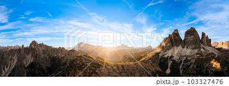 Sun sets on horizon above rocky mountain range. Sand-covered slopes of foothills of Tre Cime di Lavaredo silhouette at sunset aerial view in back lit 103327476