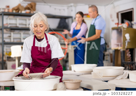 Mature woman learning how to create pottery on potter wheel in workshop Mature woman learning how to create pottery on potter wheel in workshop 103328227