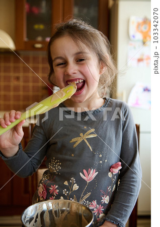 Cute little girl helps her mom in kitchen to prepare cream for the cake, quietly licks a spoon with melted delicious confectionery chocolate, mischievously smiling and making faces looking at camera 103342070