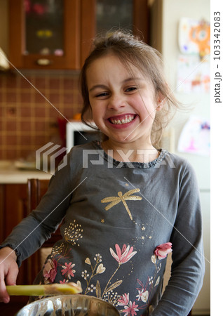 Adorable mischievous Caucasian little child girl making faces, grimacing looking at camera, tasting sweet cream whith melted Belgian chocolate, standing at the kitchen table at cozy home interior 103342083