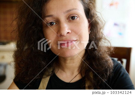 Headshot Beautiful curly haired multi ethnic woman housewife, baker confectioner in chef's apron, sitting at table in home kitchen, looking confidently at camera. Culinary. Cookery. People and hobby Headshot Beautiful curly haired multi ethnic woman housewife, baker confectioner in chef's apron, sitting at table in home kitchen, looking confidently at camera. Culinary. Cookery. People and hobby 103342124