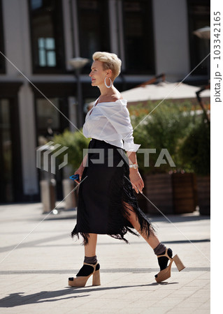 Confidence and sophistication mature woman walks city streets in fashionable ensemble chic white shirt and black fringed skirt with high heeled sandals and socks, fashion icon of style Confidence and sophistication mature woman walks city streets in fashionable ensemble chic white shirt and black fringed skirt with high heeled sandals and socks, fashion icon of style 103342165