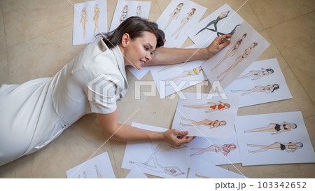 A woman lies on the floor and looks at sketches of swimwear and underwear.  103342652