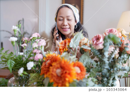 Startup, small business, flower shop. Older florist putting a flower on a vase and talking with customer by phone 103343064