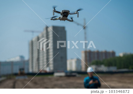 A man in a helmet and overalls controls a drone at a construction site. The builder carries out technical oversight. A man in a helmet and overalls controls a drone at a construction site. The builder carries out technical oversight. 103346872
