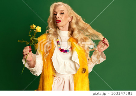 Portrait of beautiful lady with long blonde hair and makeup, senior woman in stylish clothes holding yellow roses against green studio background Portrait of beautiful lady with long blonde hair and makeup, senior woman in stylish clothes holding yellow roses against green studio background 103347393