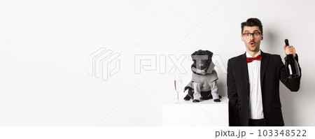 Pets, winter holidays and New Year concept. Cheerful man with cute black pug dog celebrating Christmas party, holding champagne bottle and smiling, white background Pets, winter holidays and New Year concept. Cheerful man with cute black pug dog celebrating Christmas party, holding champagne bottle and smiling, white background 103348522
