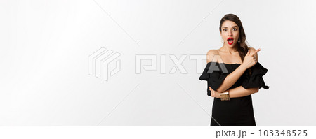 Fashion and beauty. Beautiful woman in black dress, red lips, pointing finger right at promo offer, looking amazed at camera, white background 103348525