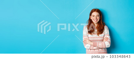 Portrait of happy redhead girl in winter sweater, smiling with arms crossed on chest, standing against blue background 103348643
