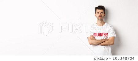 Serious young male volunteer in white t-shirt, holding arms crossed on chest, looking at camera, ready to help Serious young male volunteer in white t-shirt, holding arms crossed on chest, looking at camera, ready to help 103348704