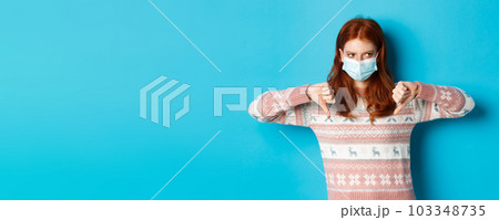 Winter, covid-19 and pandemic concept. Moody redhead girl in face mask showing thumbs-down, disapprove and dislike, standing against blue background Winter, covid-19 and pandemic concept. Moody redhead girl in face mask showing thumbs-down, disapprove and dislike, standing against blue background 103348735