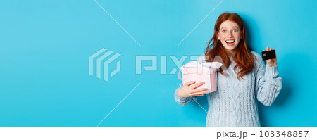 Winter holidays promo offer concept. Cheerful redhead woman holding Christmas gift and credit card, staring at camera amazed, standing over blue background 103348857