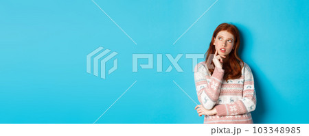 Image of thoughtful redhead girl looking at upper left corner, thinking or making choice, standing in winter sweater over blue background Image of thoughtful redhead girl looking at upper left corner, thinking or making choice, standing in winter sweater over blue background 103348985