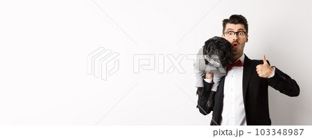 Happy young hipster in suit and glasses, showing thumb-up, holding cute black dog on shoulder, love his pug, standing over white background Happy young hipster in suit and glasses, showing thumb-up, holding cute black dog on shoulder, love his pug, standing over white background 103348987