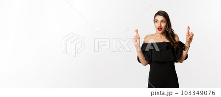 Fashion and beauty. Excited gorgeous woman in black dress, holding fingers crossed and looking at upper left corner, making wish, standing over white background 103349076