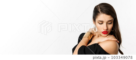 Fashion and beauty concept. Close-up of sensual woman in elegant earrings and black dress, wearing makeup with red lips, looking down tenderly, standing over white background 103349078