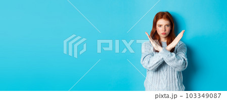 Serious redhead girl looking confident, showing cross gesture to stop and forbid action, standing over blue background Serious redhead girl looking confident, showing cross gesture to stop and forbid action, standing over blue background 103349087
