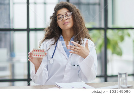 Young pharmacist woman holding pills while sitting at the table in the office Young pharmacist woman holding pills while sitting at the table in the office 103349090