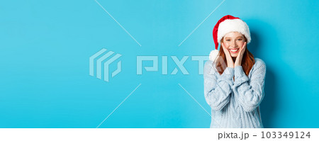 Winter holidays and Christmas Eve concept. Cheerful redhead girl in santa hat, celebrating New Year, blushing and smiling happy, standing over blue background Winter holidays and Christmas Eve concept. Cheerful redhead girl in santa hat, celebrating New Year, blushing and smiling happy, standing over blue background 103349124