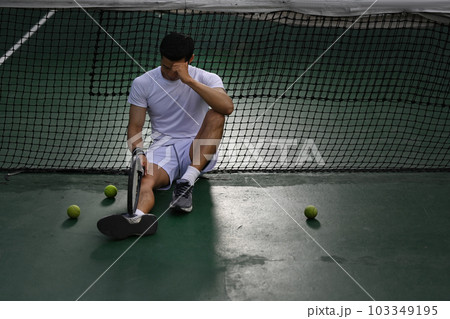 Disappointed asian male tennis player sitting on the court, upset about competition loss, failure emotions 103349195