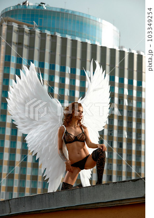 Young sexy fallen angel brunette girl in black lingerie and stockings with white wings behind her back sits in a seductive pose on the edge of the roof against the backdrop of a modern skyscraper. Young sexy fallen angel brunette girl in black lingerie and stockings with white wings behind her back sits in a seductive pose on the edge of the roof against the backdrop of a modern skyscraper. 103349254