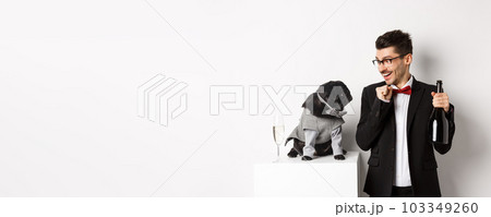 Cheerful young man in suit celebrating New Year with pet, dog and guy looking at each other, owner drinking champagne, standing over white background Cheerful young man in suit celebrating New Year with pet, dog and guy looking at each other, owner drinking champagne, standing over white background 103349260