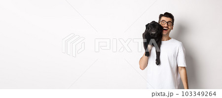 Cheerful hipster guy in glasses and t-shirt, carry cute black pug on shoulder and smiling. Dog owner playing with his pet, standing over white background Cheerful hipster guy in glasses and t-shirt, carry cute black pug on shoulder and smiling. Dog owner playing with his pet, standing over white background 103349264