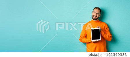 Technology. Handsome adult man showing digital tablet screen, smiling and looking at upper left corner thoughtful, imaging or daydreaming, blue background Technology. Handsome adult man showing digital tablet screen, smiling and looking at upper left corner thoughtful, imaging or daydreaming, blue background 103349388