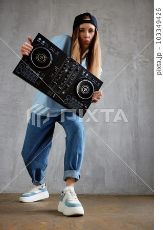 A young pretty long haired DJ girl in a blue sweater, jeans and a black baseball cap poses with a black DJ mixing console. Studio shot, gray background. A young pretty long haired DJ girl in a blue sweater, jeans and a black baseball cap poses with a black DJ mixing console. Studio shot, gray background. 103349426
