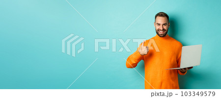 Freelance and technology concept. Lucky man in orange sweater, showing thumb up while working with laptop, standing satisfied over light blue background 103349579