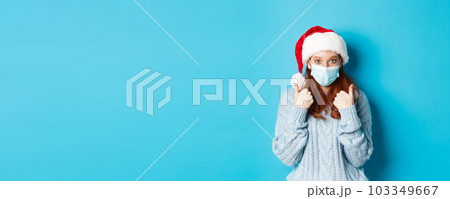 Christmas, quarantine and covid-19 concept. Cute teen redhead girl in santa hat and sweater, wearing face mask from coronavirus, showing thumbs up, standing over blue background Christmas, quarantine and covid-19 concept. Cute teen redhead girl in santa hat and sweater, wearing face mask from coronavirus, showing thumbs up, standing over blue background 103349667