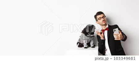 Cheerful dog owner in suit celebrating New Year with dog, taking selfie on smartphone near cute black pug in costume, white background 103349681