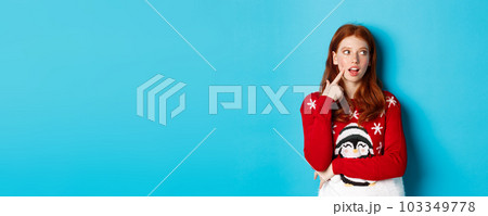 Winter holidays and Christmas Eve concept. Pretty redhead girl in xmas sweater, touching cheek thoughtful and smiling, making choice, looking at upper right corner and thinking, blue background 103349778