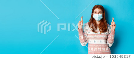 Winter, coronavirus and social distancing concept. Cute hopeful girl with red hair, wearing face mask, cross fingers and making wish, standing over blue background 103349817