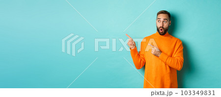 Surprised and curious guy looking at promo, pointing at upper left corner copy space, standing in orange sweater, turquoise background 103349831